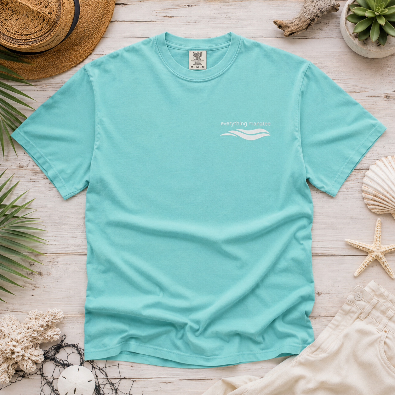A mint green short-sleeve t-shirt with a white wave graphic and 'Everything Matters' text on the left chest, styled in a flat lay with a straw hat, seashells, starfish, and palm leaves on a white wood background.