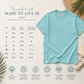 A light blue short-sleeve t-shirt displayed on a cream background with a size chart, fit text, and feature icons. The shirt is styled with beach-themed props including a woven hat, seashell, and palm leaves.