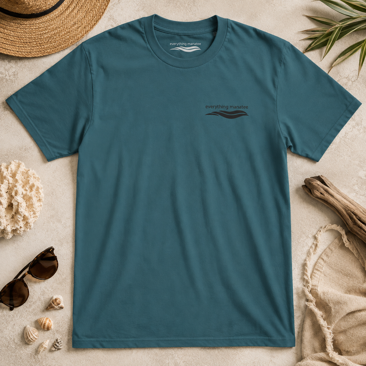 A teal blue short-sleeve t-shirt with a small dark graphic on the left chest reading 'Everything Supplies' and a wave design, laid flat on a light beige textured surface with beach-themed props including a straw hat, sunglasses, shells, driftwood, and a towel.