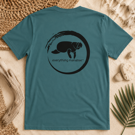 A teal blue short-sleeve t-shirt laid flat on a sandy surface with beach-themed props like driftwood, a woven mat, and a palm leaf. The back of the shirt features a black circular graphic with a manatee silhouette and the text 'everything manatee'.