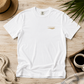 A white short-sleeve crewneck t-shirt with a small gold logo on the left chest, laid flat on a light wood surface with a straw hat, rope, coffee cup, and green leaves as styled props.