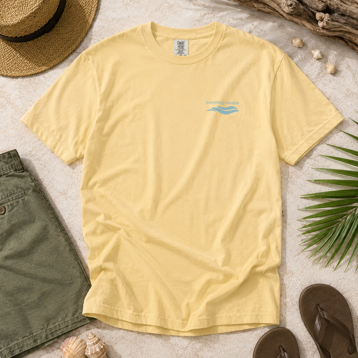 A pale yellow short-sleeve t-shirt with a small light blue wave graphic and 'The Watermark' brand name on the left chest, styled in a flat lay with a straw hat, green shorts, seashells, a palm leaf, and brown flip-flops on a light sandy background.