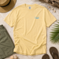A pale yellow short-sleeve t-shirt with a small light blue wave graphic and 'The Watermark' brand name on the left chest, styled in a flat lay with a straw hat, green shorts, seashells, a palm leaf, and brown flip-flops on a light sandy background.