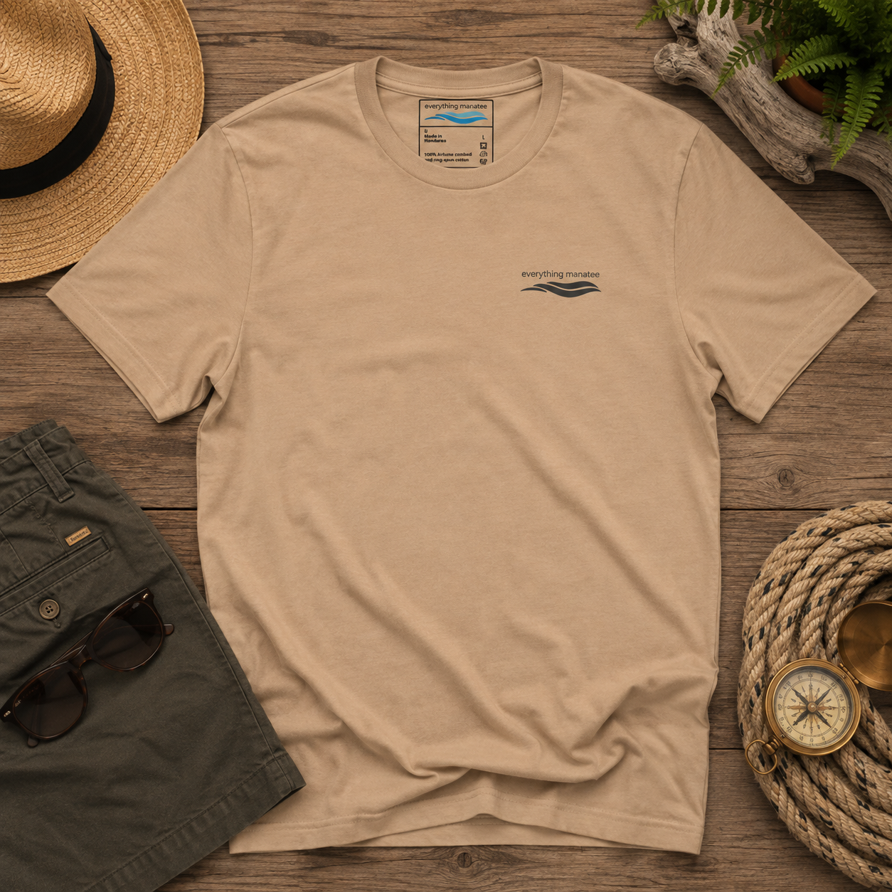 A beige short-sleeve t-shirt with a small dark blue wave graphic on the left chest, laid flat on a rustic wooden surface surrounded by a straw hat, green shorts, sunglasses, a compass, and a potted fern.