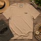 A beige short-sleeve t-shirt with a small dark blue wave graphic on the left chest, laid flat on a rustic wooden surface surrounded by a straw hat, green shorts, sunglasses, a compass, and a potted fern.