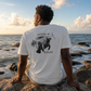 A man sits on rocks by the ocean, viewed from behind, wearing a white graphic T-shirt with a black illustration of a sea lion and the words 'Inhale' and 'Exhale'.