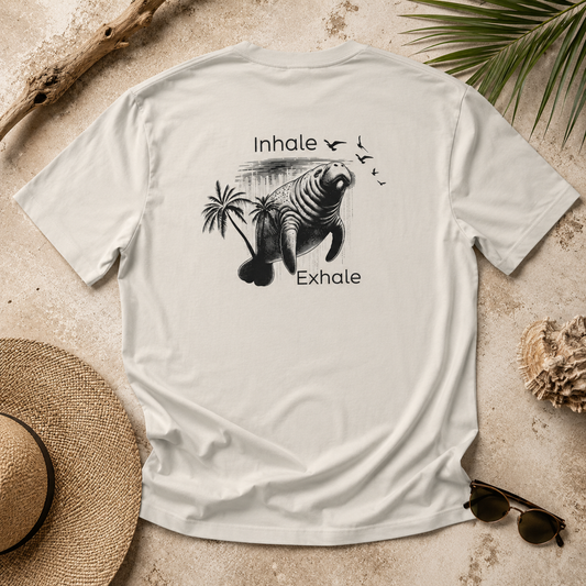 A light beige crewneck t-shirt laid flat on a sandy surface with beach-themed props including a straw hat, sunglasses, driftwood, and palm leaves. The back of the shirt features a black graphic of a manatee with palm trees and the words 'Inhale' and 'Exhale'.