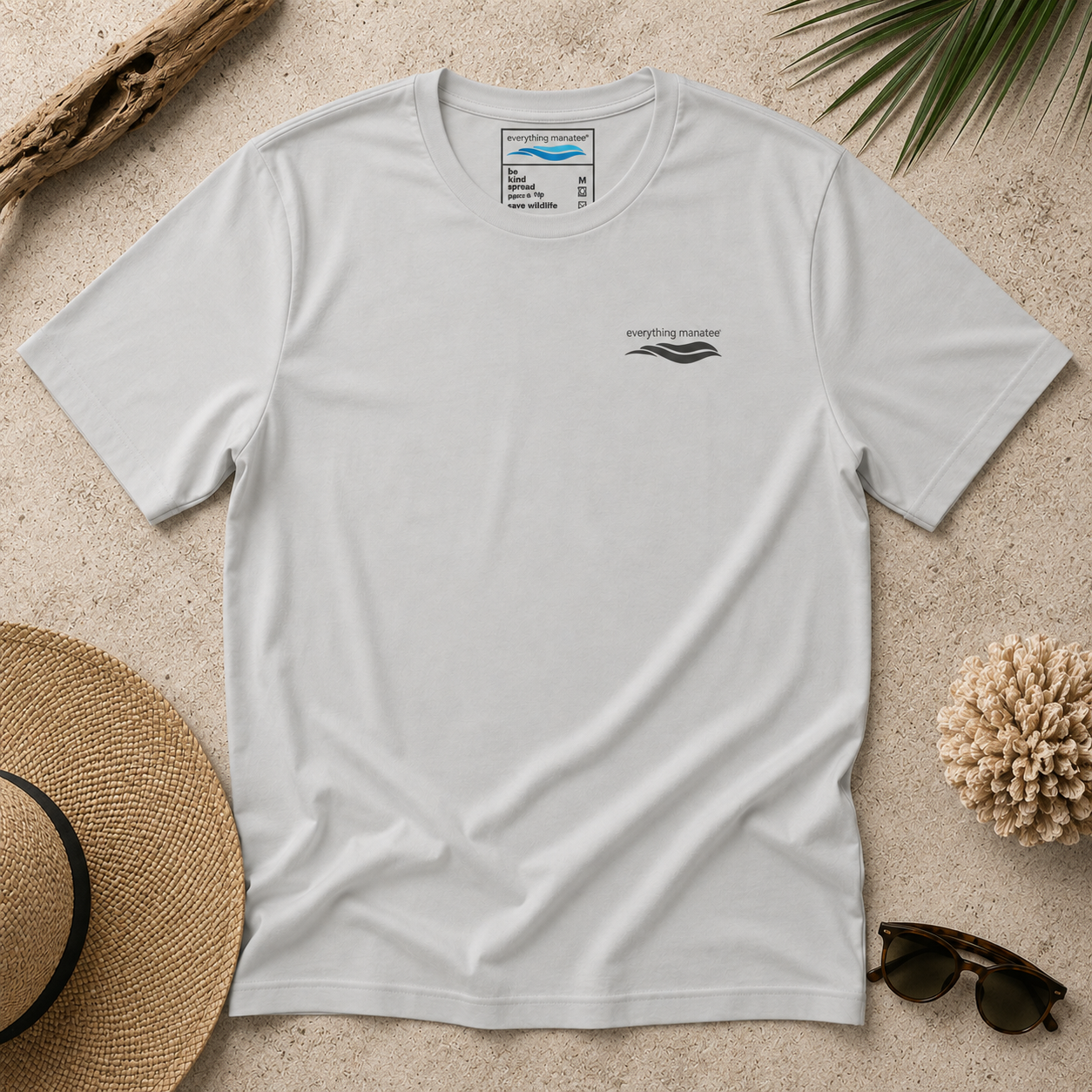 A light gray short-sleeve t-shirt with a small black chest graphic, laid flat on a sandy background with a straw hat, sunglasses, driftwood, and palm leaves as props.