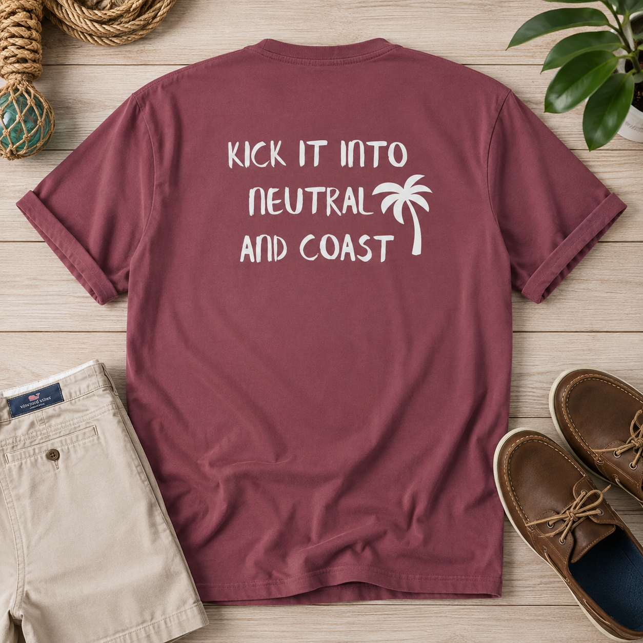 A maroon short-sleeve t-shirt is laid flat on a light wood surface, showing the back with white text that reads 'KICK IT INTO NEUTRAL AND COAST' alongside a white palm tree graphic. Beige shorts, brown boat shoes, rope, and a green plant are styled around it as props.