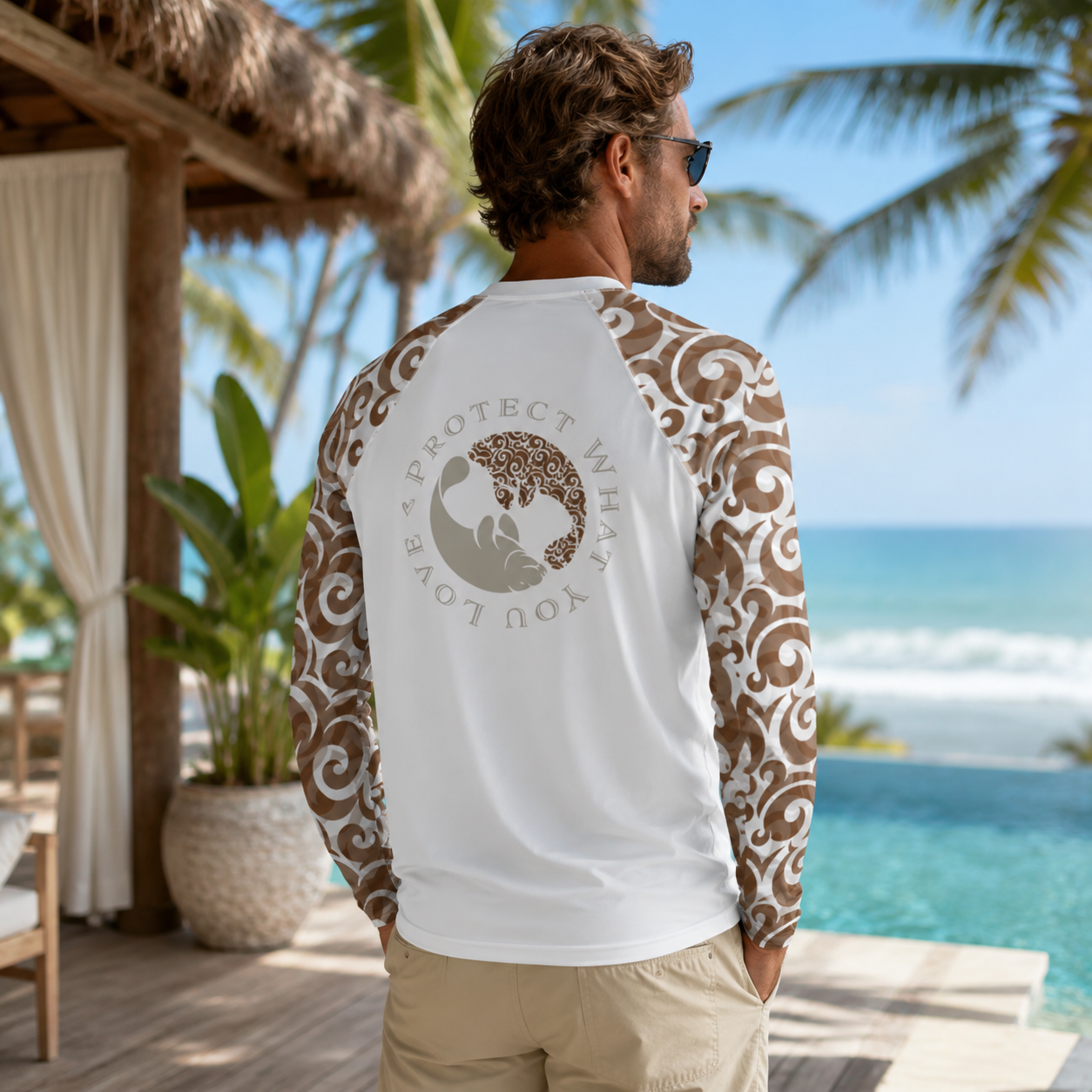 A man wearing a white long-sleeve rash guard with brown patterned sleeves and a large turtle graphic on the back, standing on a tropical beach deck with the ocean in the background.