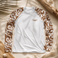 A long-sleeve raglan shirt laid flat on a beige fabric surface with decorative palm leaves, driftwood, and woven straw. The shirt is white with brown and white scroll-patterned sleeves and a small chest logo reading 'everything natural™'.