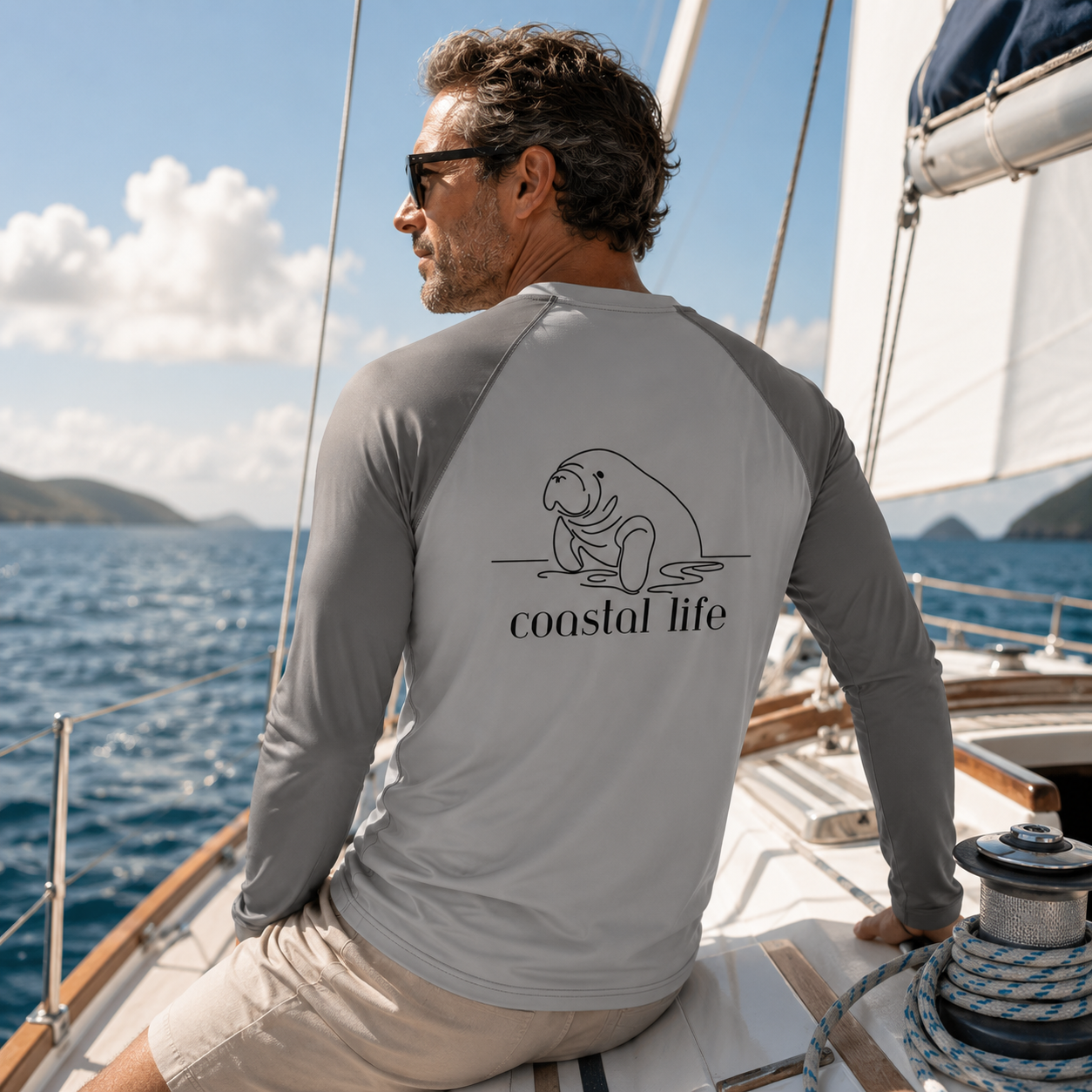 A man wearing a gray long-sleeve rash guard with a black line art seal and the text 'coastal life' on the back, sitting on a sailboat deck with the ocean and sky in the background.