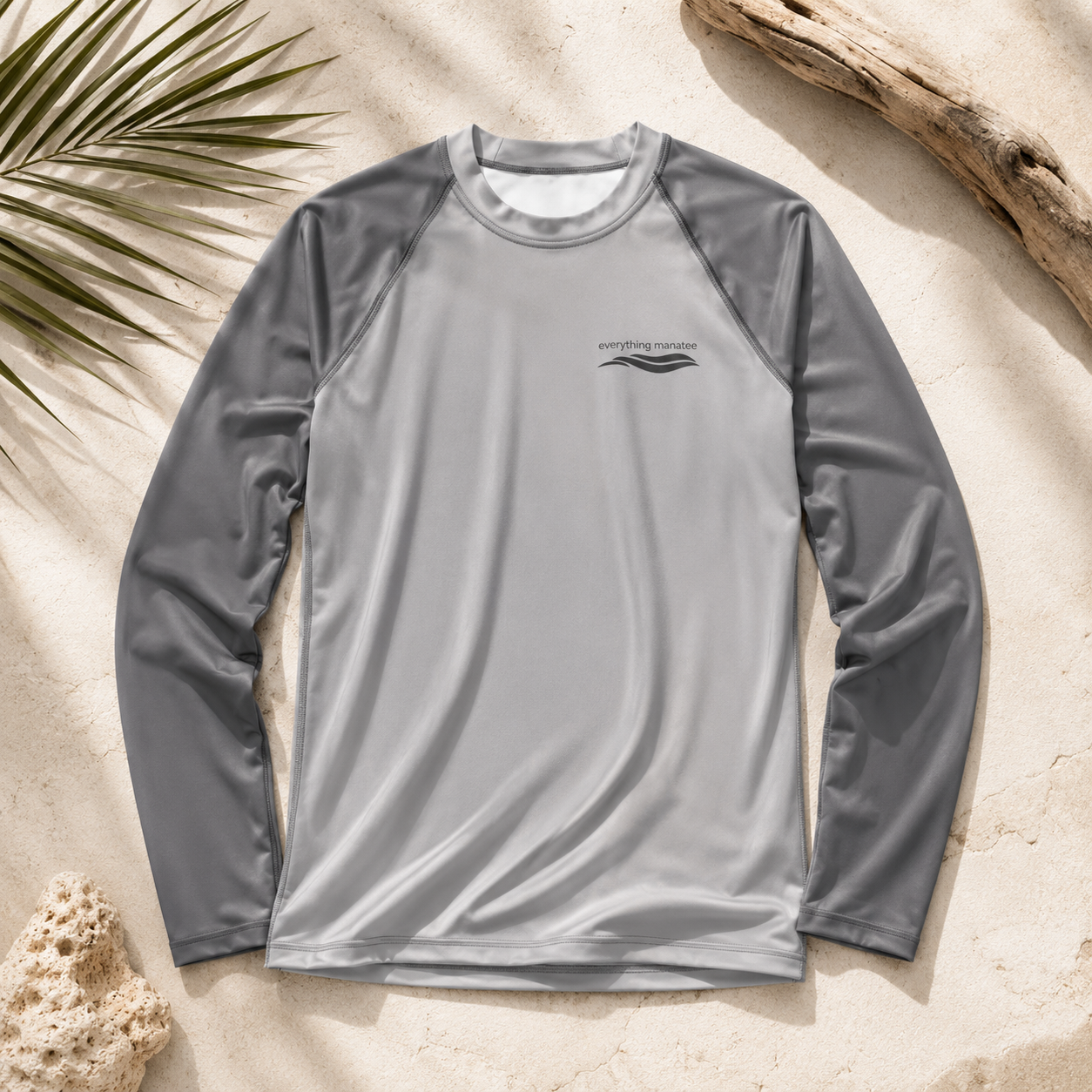 A long-sleeve athletic rash guard in light gray with darker gray raglan sleeves, laid flat on a sandy surface with beach-themed props like palm leaves, driftwood, and coral.