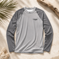 A long-sleeve athletic rash guard in light gray with darker gray raglan sleeves, laid flat on a sandy surface with beach-themed props like palm leaves, driftwood, and coral.