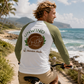 A man riding a bicycle on a beach path wearing a white and olive green raglan long-sleeve shirt with a large back graphic that reads 'UNWIND' and 'Manatee Mode'.