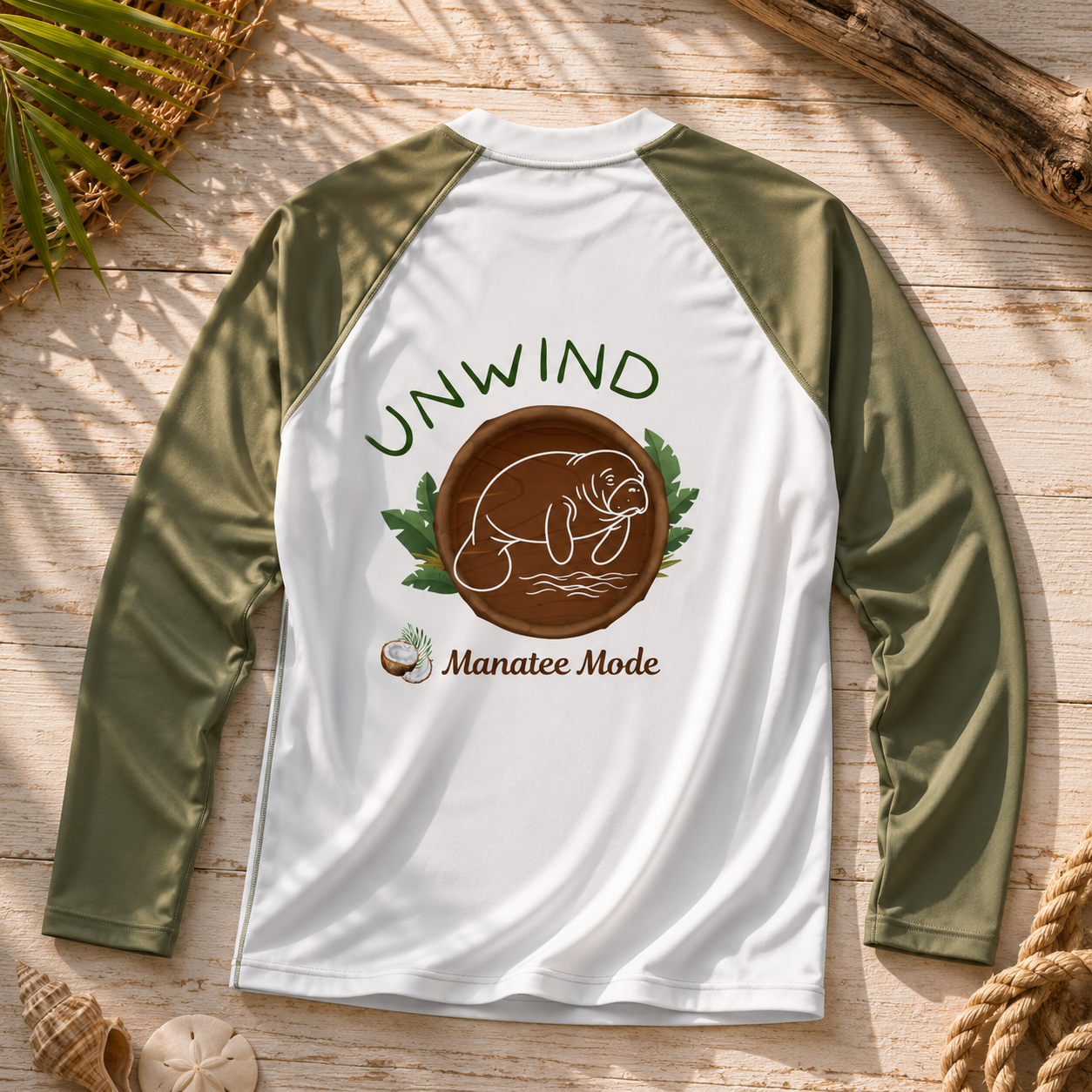 A long-sleeve raglan shirt laid flat on a rustic wood surface with seashell and rope props. The shirt has an olive green body and sleeves with a white collar, featuring a large back graphic that reads “UNWIND” above a brown seal illustration and the words “Manatee Mode.