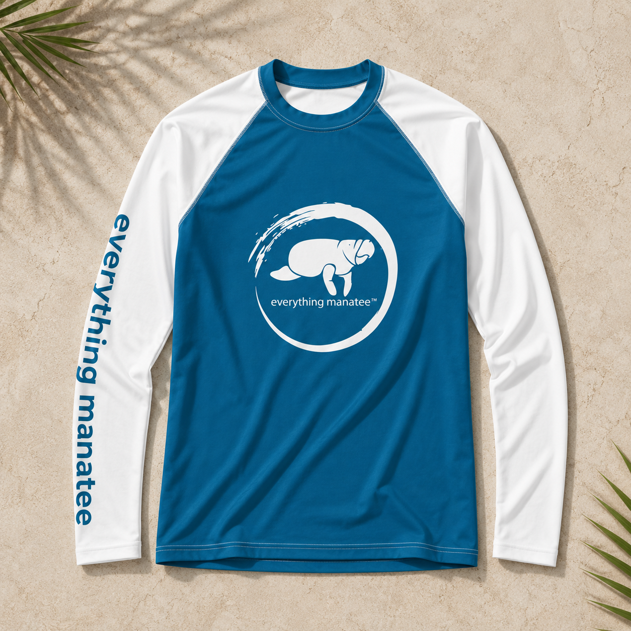 A long-sleeve raglan shirt laid flat on a textured beige surface with palm leaves in the corners. The shirt is blue on the torso and sleeves, with white sleeves and a white circular turtle logo on the chest.