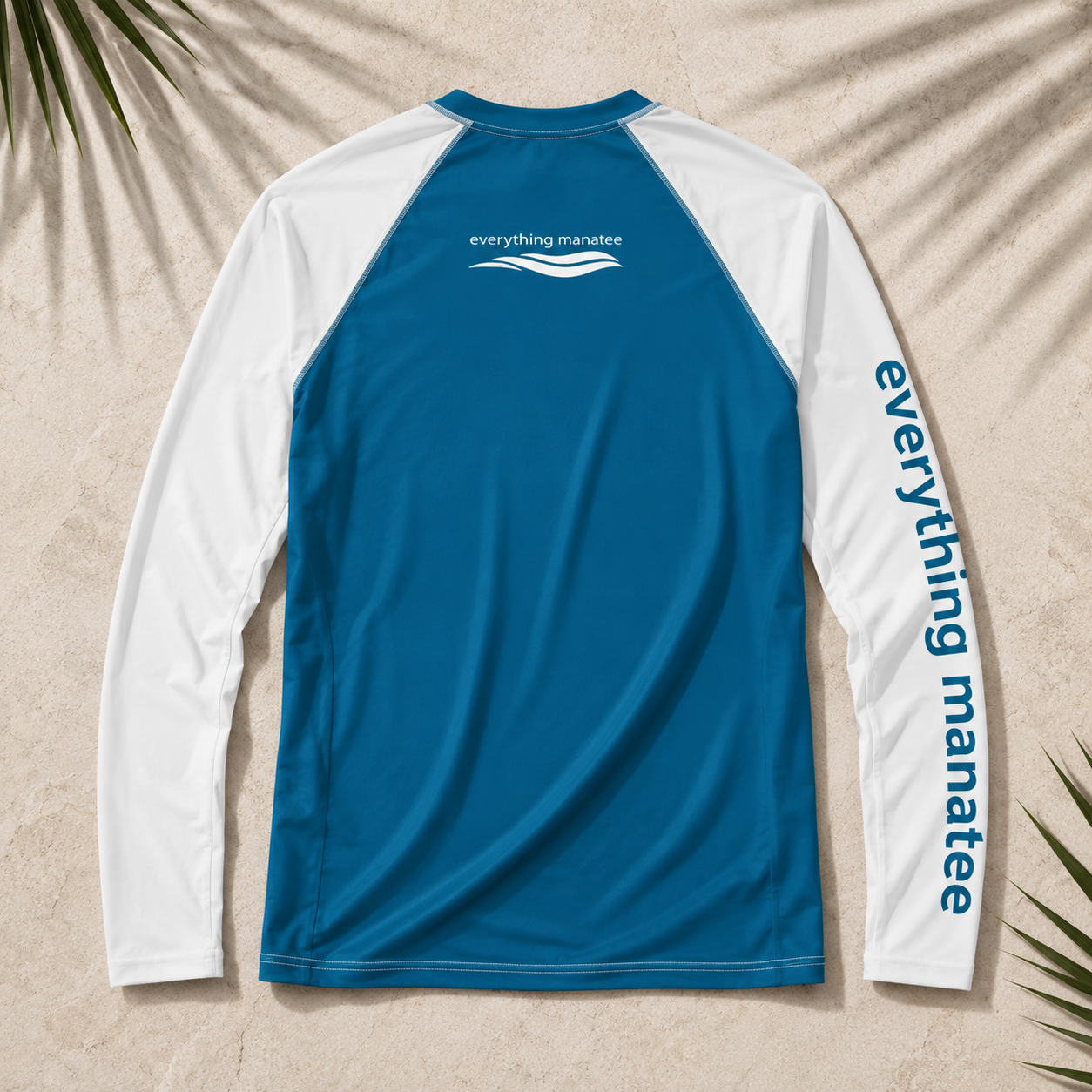 A long-sleeve raglan performance top laid flat on a sandy surface with palm leaf shadows. The back is bright blue, the sleeves and shoulder yoke are white, and the brand name 'everything manatee' is printed in white on the upper back and down the right sleeve.