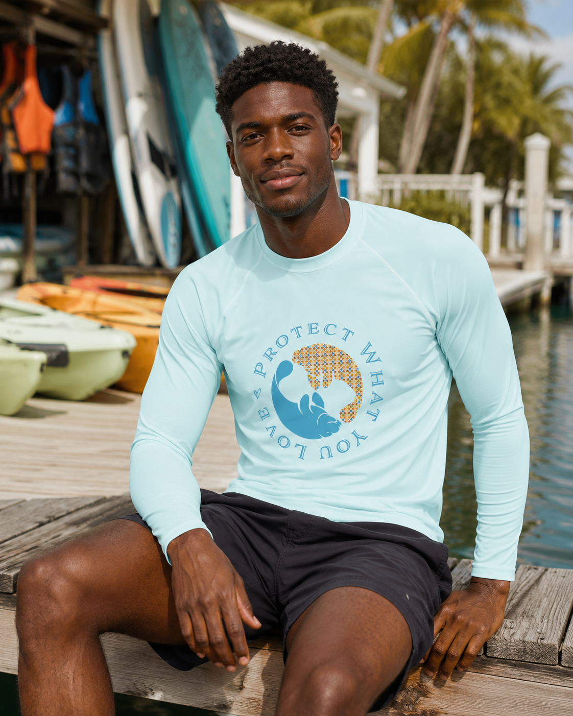 A man sitting on a wooden dock wearing a light blue long-sleeve rash guard with a blue and gold graphic on the chest that reads 'PROTECT WHAT YOU LOVE'.