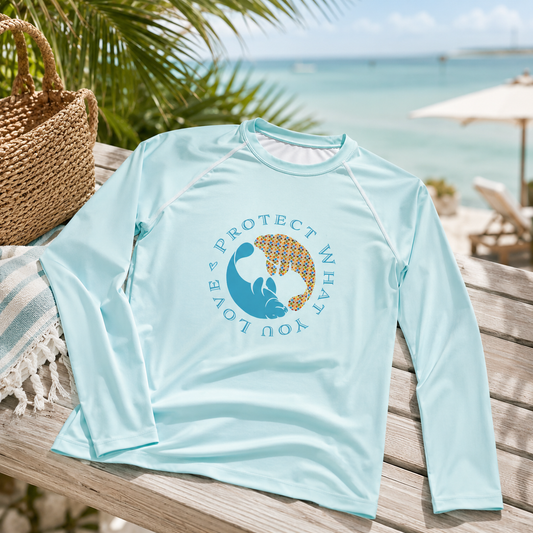 A light blue long-sleeve rash guard shirt with a graphic of a manatee and the text 'PROTECT WHAT YOU LOVE' laid out on a wooden beach chair with a straw hat and tropical beach background.