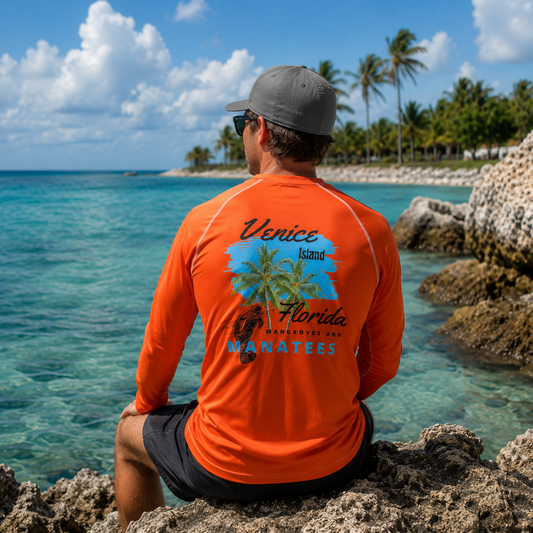 A man wearing a bright orange long-sleeve rash guard with a 'Venice Island Florida Manatees' graphic on the back, sitting on a rocky beach with turquoise water and palm trees in the background.