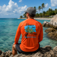 A man wearing a bright orange long-sleeve rash guard with a 'Venice Island Florida Manatees' graphic on the back, sitting on a rocky beach with turquoise water and palm trees in the background.