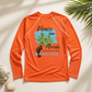 An orange long-sleeve rash guard shirt laid flat on a light beige background with decorative seashells and palm leaves. The shirt features a graphic print on the front with a blue sky, green palm tree, a stingray, and text reading 'Venice Island Florida Mangroves and Manatees'.