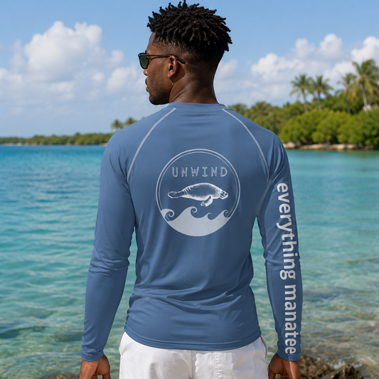 A man stands on a beach with turquoise water and palm trees in the background, wearing a blue long-sleeve rash guard from the brand Unwind. The back of the shirt features a white circular wave graphic with the brand name 'UNWIND' and a fish, while the left sleeve has the phrase 'everything manatees' printed vertically in white.