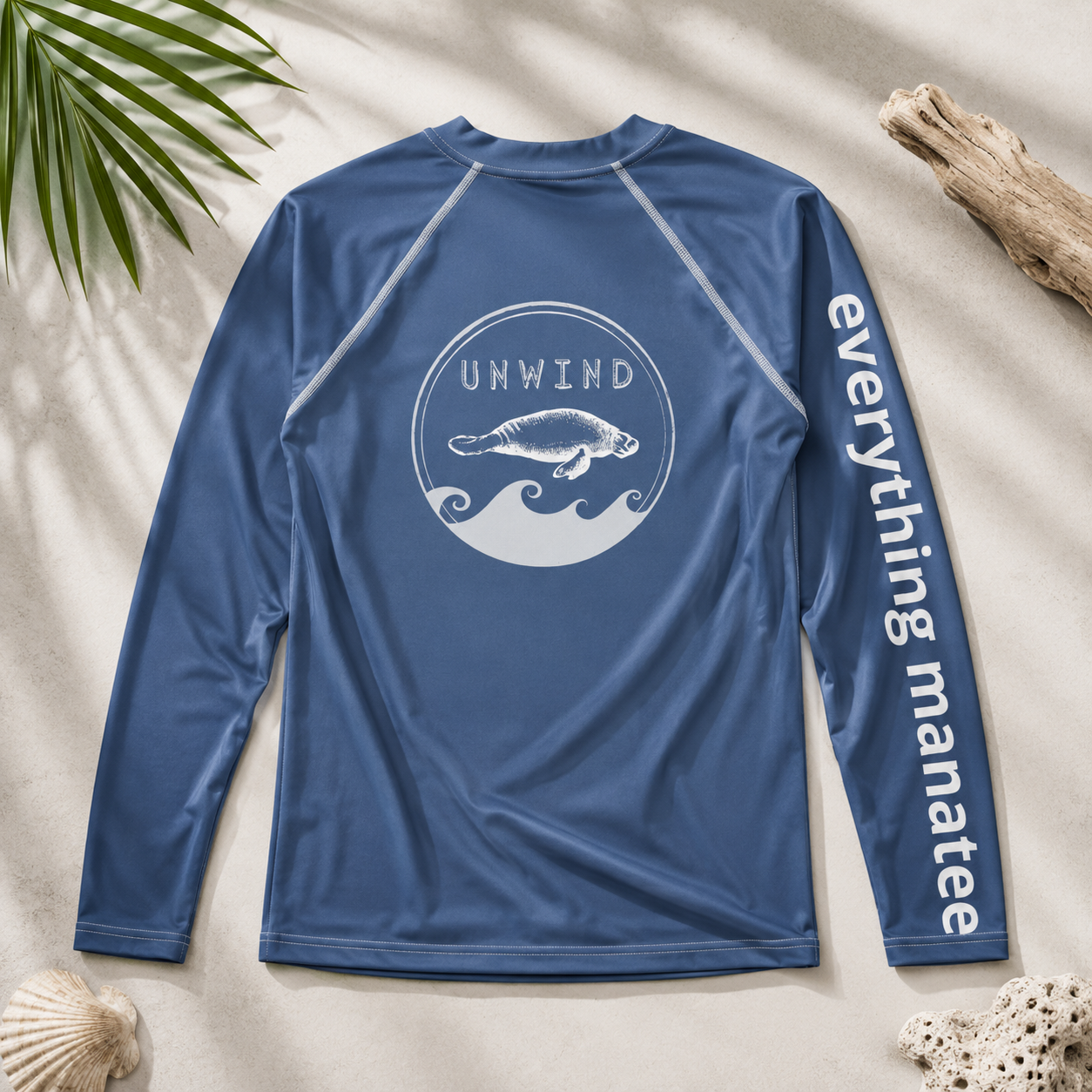 A blue long-sleeve rash guard shirt laid flat on a sandy surface with beach props. The back has a white circular wave graphic with a manatee and the word 'UNWIND' above it, and the right sleeve has 'everything manatee' printed vertically in white.