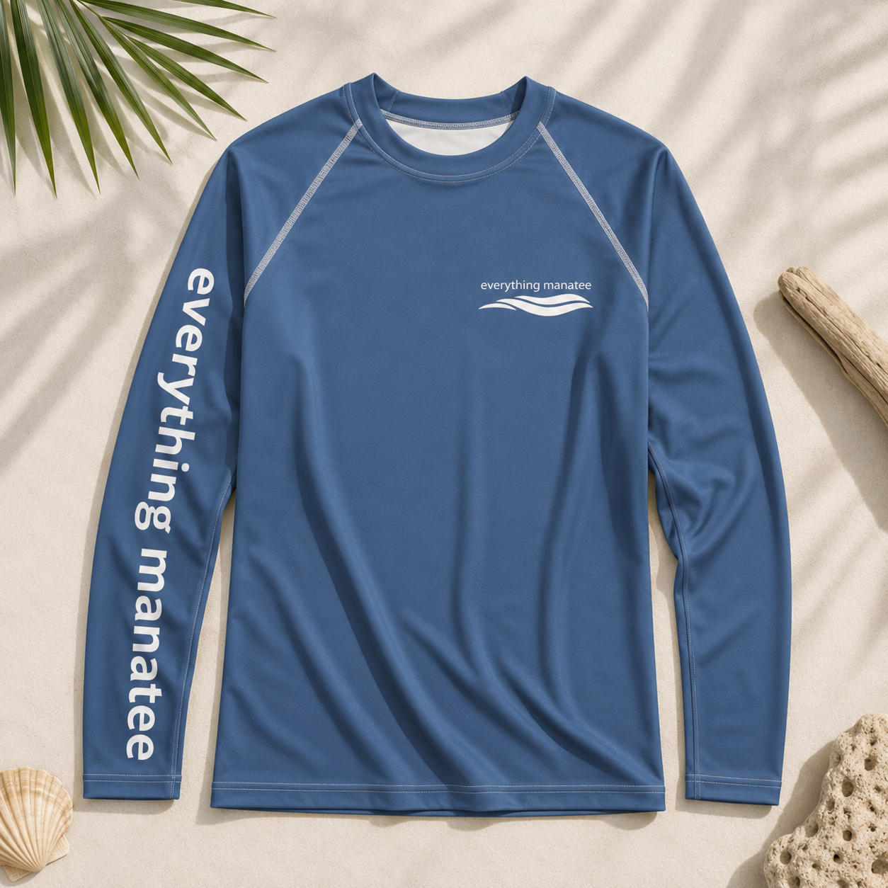 A blue long-sleeve rash guard with 'everything manatee' printed in white down the left sleeve and a small white chest logo, laid flat on a sandy background with beach-themed props.