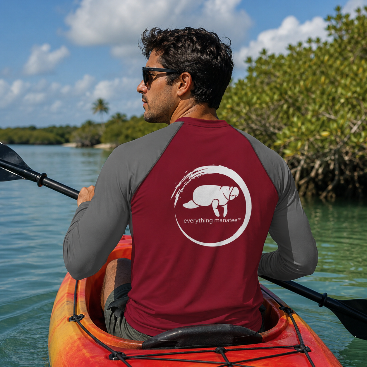 A man wearing sunglasses sits in an orange kayak on the water, viewed from behind. He wears a long-sleeve rash guard with a dark red back panel and gray sleeves, featuring a white circular logo with a manatee and the text 'everything marinet'.