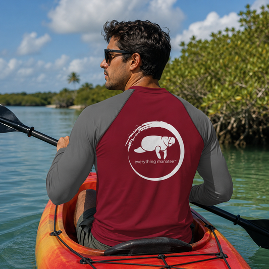 A man wearing sunglasses sits in an orange kayak on the water, viewed from behind. He wears a long-sleeve rash guard with a dark red back panel and gray sleeves, featuring a white circular logo with a manatee and the text 'everything marinet'.