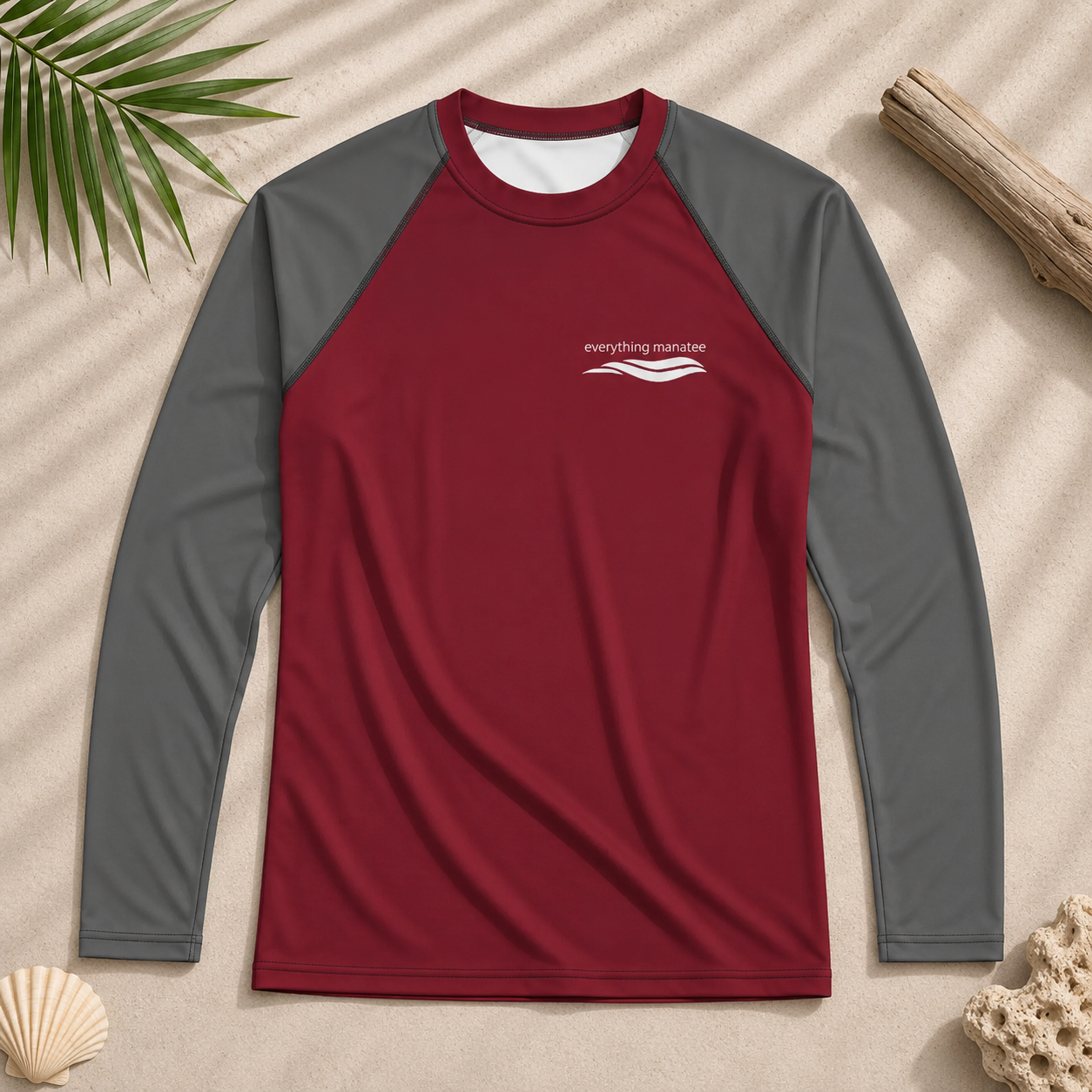A long-sleeve raglan rash guard shirt in red and gray, laid flat on a sandy background with beach-themed props like a palm leaf, driftwood, and a seashell.
