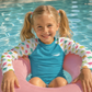 A young girl with blonde pigtails sits in a pink inflatable pool float in a swimming pool, wearing a blue and white long-sleeve swim shirt with a colorful dinosaur print on the sleeves.
