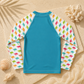 A long-sleeve rash guard swimsuit laid flat on a sandy background with seashells and netting. The back of the shirt is solid turquoise blue, and the sleeves have a white background patterned with small colorful dinosaur silhouettes in pink, orange, green, and blue.
