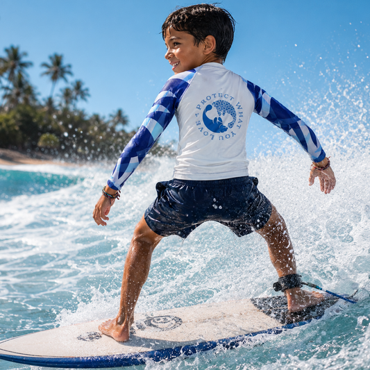 A young boy surfing on a wave while wearing a white and blue long-sleeve rash guard with a 'Surf Life International' logo on the back, dark swim trunks, and a black ankle leash on a white surfboard.