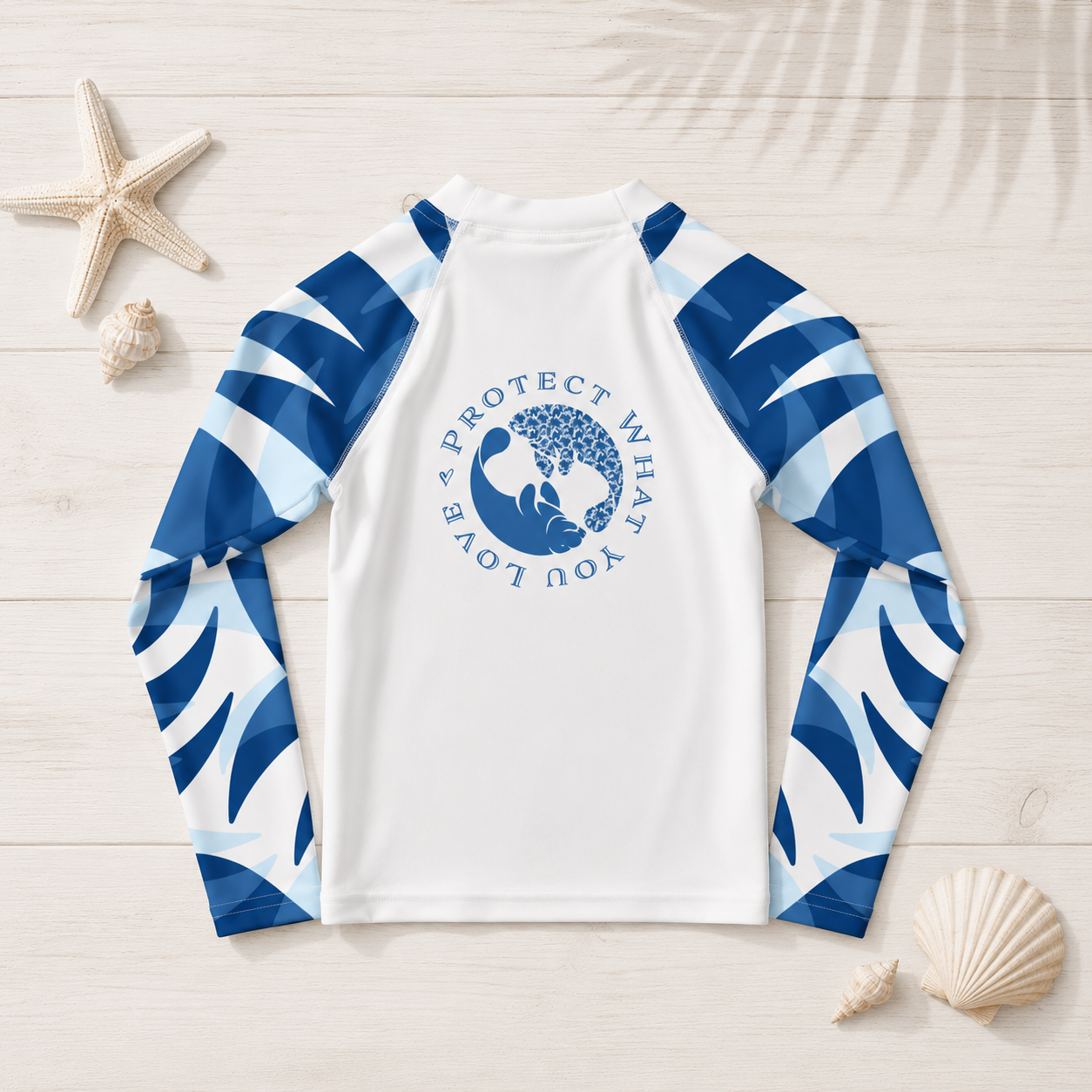 A long-sleeve raglan rash guard laid flat on a light wood background with seashell and starfish props. The shirt is white with blue and white wave-patterned sleeves and a blue circular whale graphic on the chest reading 'A PROTECT WHAT YOU LOVE'.