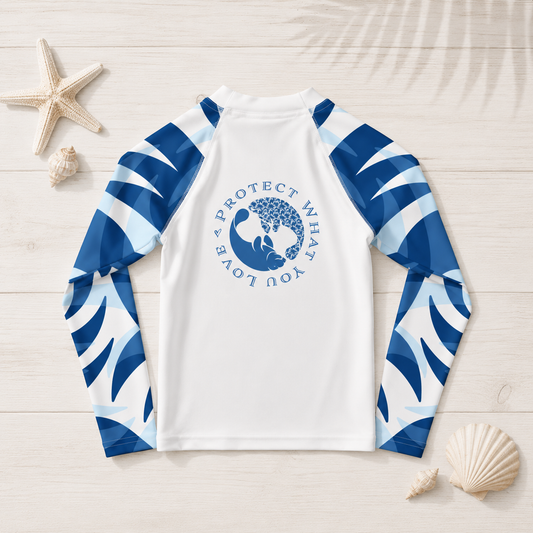 A long-sleeve raglan rash guard laid flat on a light wood background with seashell and starfish props. The shirt is white with blue and white wave-patterned sleeves and a blue circular whale graphic on the chest reading 'A PROTECT WHAT YOU LOVE'.