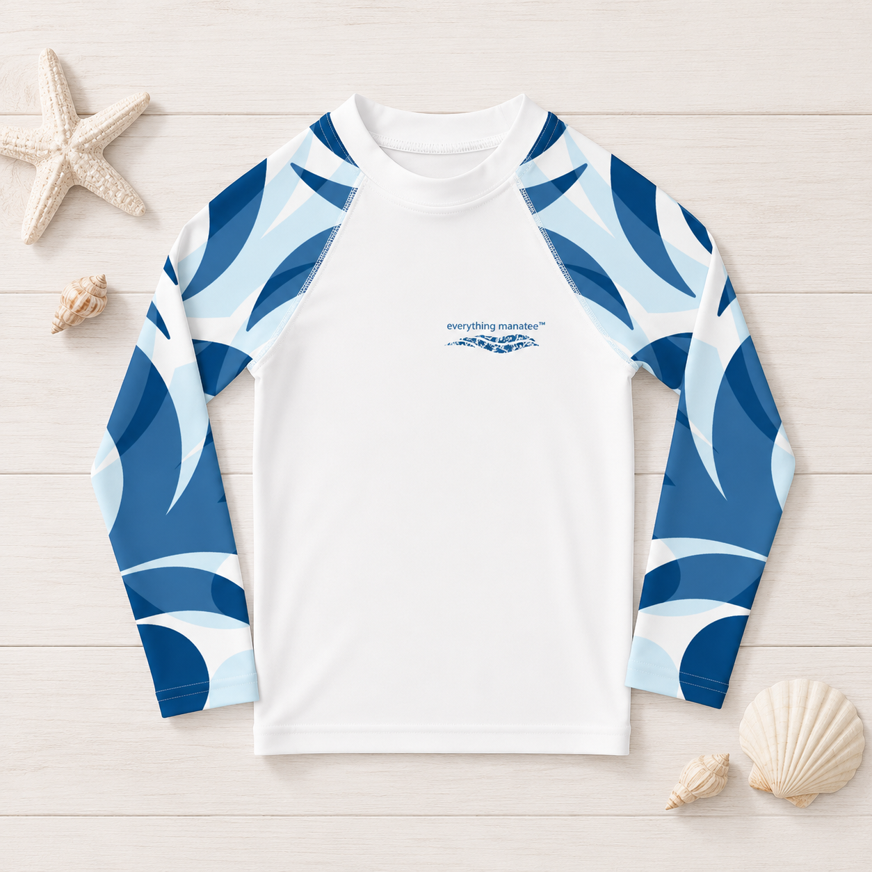 A long-sleeve raglan shirt with a white body and blue and light blue wave-patterned sleeves, laid flat on a light wood surface with seashells and a starfish around it. A small 'everything-handles' logo is printed on the chest.