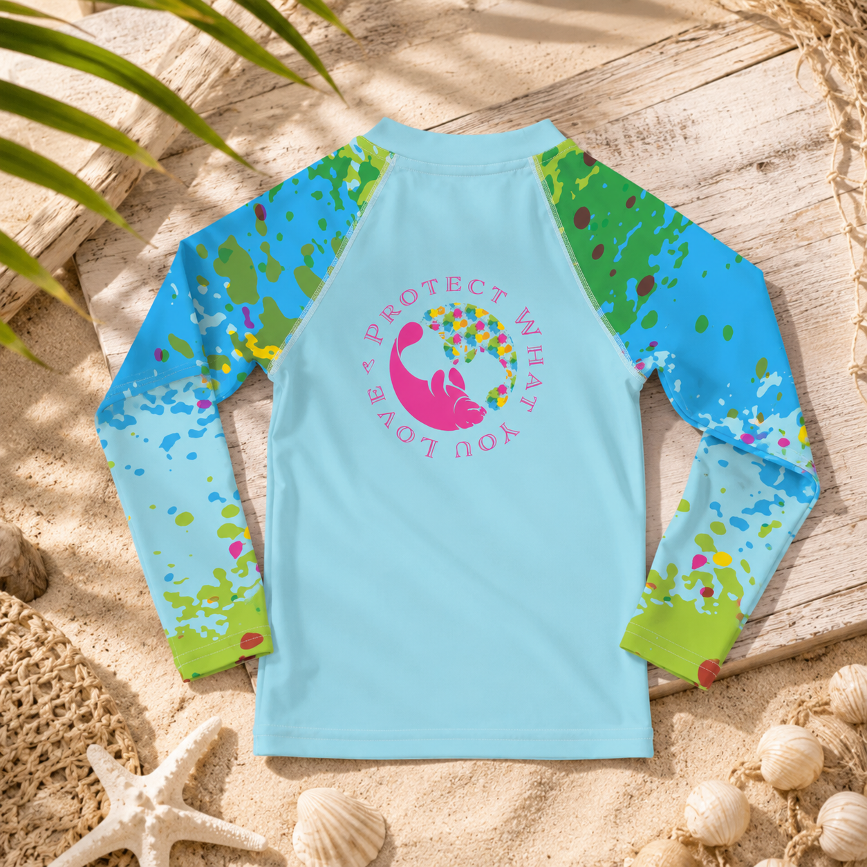 A long-sleeve kids' rash guard laid flat on a sandy beach-style surface with palm leaves, shells, and a starfish. The shirt is light blue with bright blue, green, and multicolor paint-splatter sleeves and a pink whale graphic with the words 'PROTECT WHAT YOU LOVE' on the chest.