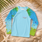 A long-sleeve kids' rash guard laid flat on a sandy surface with beach props like shells, driftwood, and palm leaves. The shirt is light blue with green and blue paint-splatter graphics on the shoulders and sleeves, and a small multicolor logo on the chest.