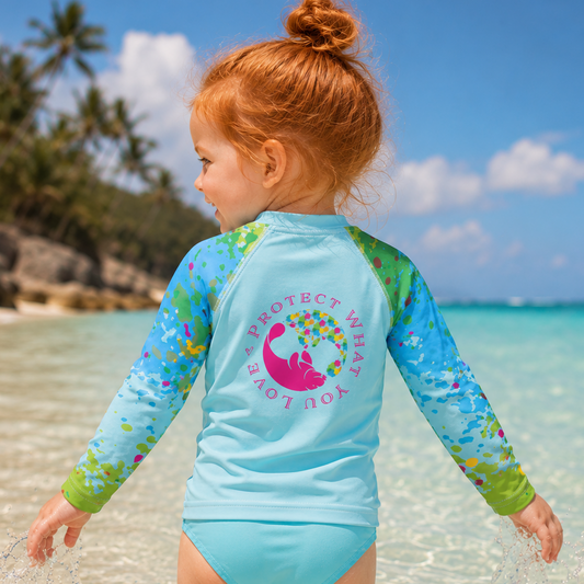 A young child standing in shallow ocean water at a beach, wearing a long-sleeve swim rash guard and matching swim bottoms. The rash guard is light blue with green and multicolor floral patterns on the sleeves and back, featuring a pink turtle graphic and the text 'PROTECT WHAT YOU LOVE.