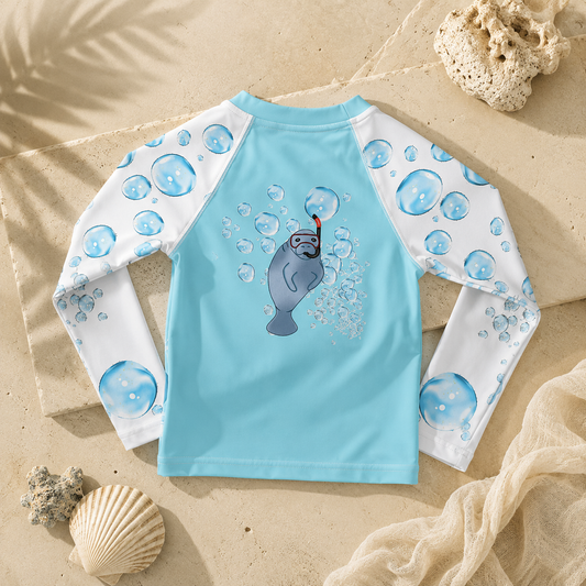 A children's long-sleeve swim shirt laid flat on a sandy surface with beach props. The back of the shirt is light blue with white sleeves printed in blue bubble patterns, and a gray manatee wearing a red snorkel graphic with bubbles on the center back.