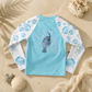 A children's long-sleeve swim shirt laid flat on a sandy surface with beach props. The back of the shirt is light blue with white sleeves printed in blue bubble patterns, and a gray manatee wearing a red snorkel graphic with bubbles on the center back.