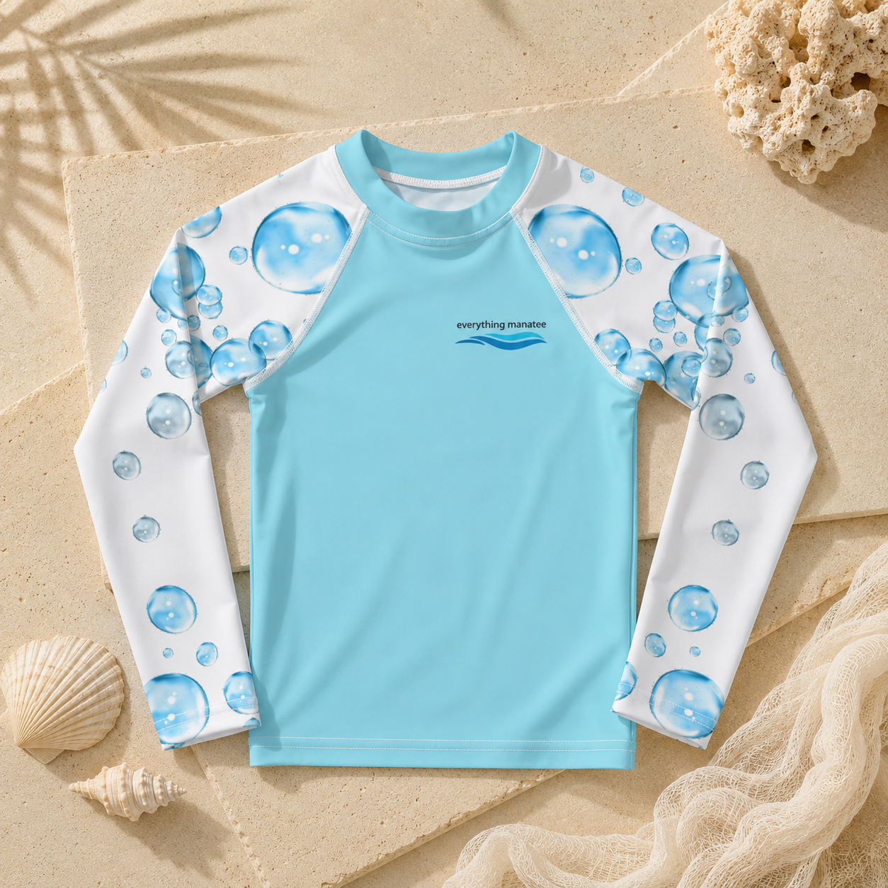 A children's long-sleeve rash guard laid flat on a sandy surface with seashell and coral props. The shirt is light blue on the torso with white sleeves printed in blue bubble patterns.