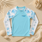 A children's long-sleeve rash guard laid flat on a sandy surface with seashell and coral props. The shirt is light blue on the torso with white sleeves printed in blue bubble patterns.