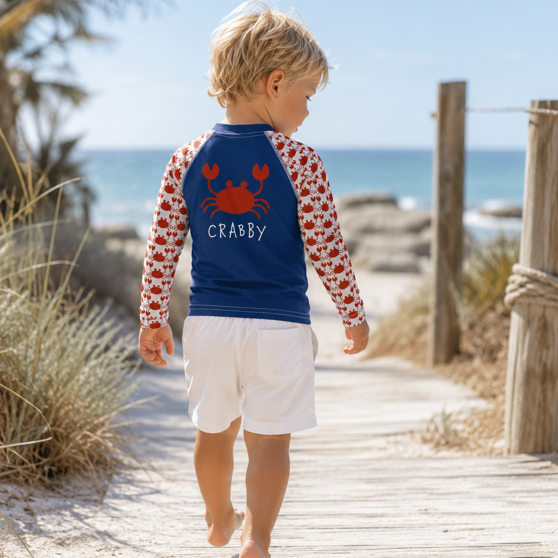 A toddler walking barefoot on a wooden boardwalk at the beach, wearing a blue and white long-sleeve rash guard with a red crab graphic and the word 'CRABBY' on the back, paired with white shorts.