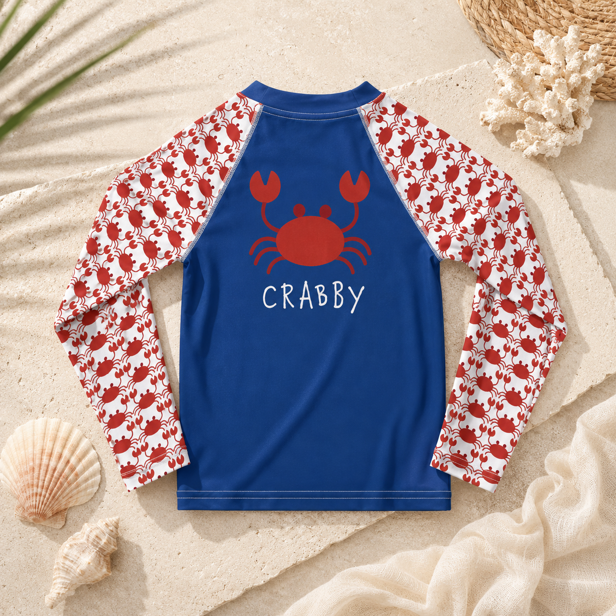 A long-sleeve children's rash guard laid flat on a sandy beach-style background with shells and woven decor. The shirt has a royal blue body with white sleeves covered in a red crab pattern, and a large red crab graphic with the word "CRABBY" printed on the back.
