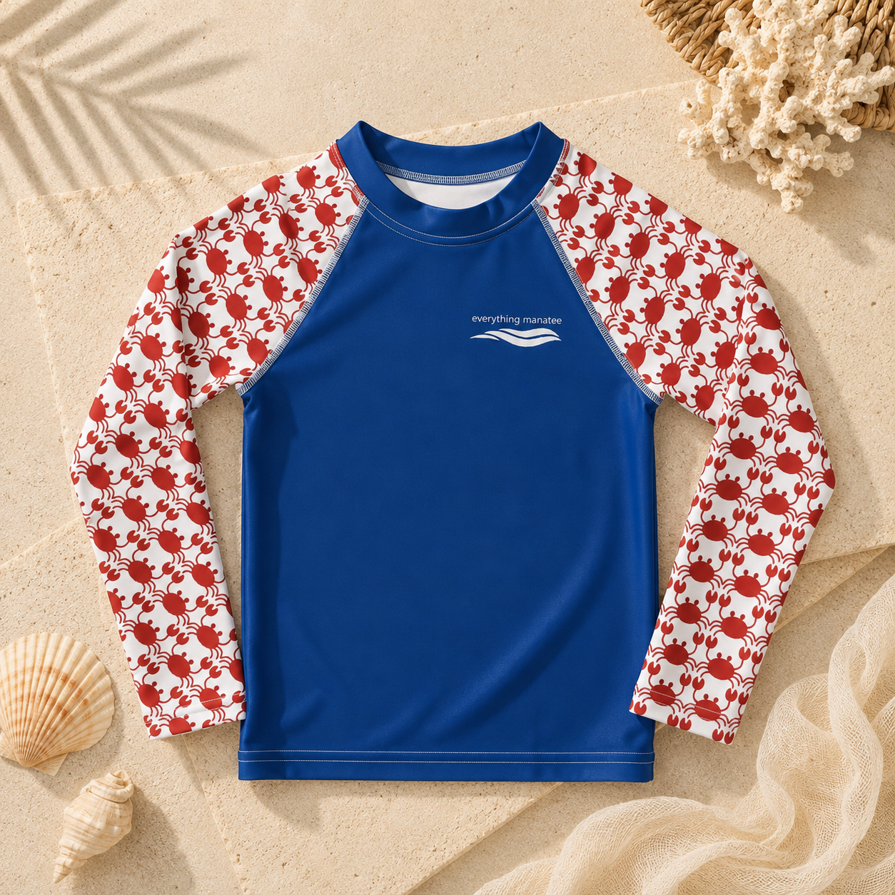 A long-sleeve kids' rash guard laid flat on a sandy surface with seashells and beach props. The shirt has a solid royal blue torso and sleeves with a red-and-white crab pattern, plus a small white 'everything marine' wave logo on the chest.