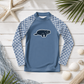 A long-sleeve kids' rash guard laid flat on a white wood background with beach-themed props. The shirt is dusty blue with a navy and white scallop pattern on the sleeves and a navy manatee graphic on the chest.